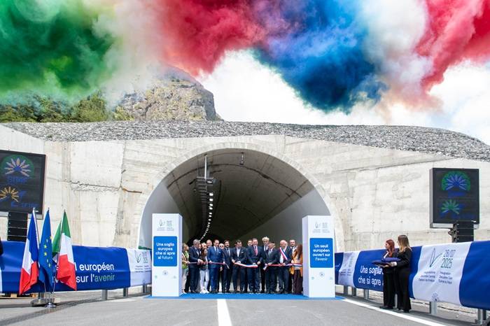 Cerimonia di inaugurazione di un tunnel con il taglio del nastro, alla presenza di numerose autorità e rappresentanti istituzionali. Sullo sfondo, fumi colorati nei colori della bandiera italiana (verde, bianco e rosso) e francese (blu, bianco e rosso) si alzano nel cielo. Ai lati dell'ingresso del tunnel sono presenti pannelli con scritte in francese e italiano che celebrano l'apertura del passaggio come 'una soglia che si apre al futuro'. Bandiere italiane, francesi ed europee decorano la scena