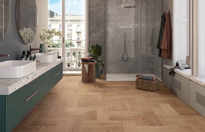 Bagno moderno e luminoso con pavimento effetto legno posato a spina di pesce. A sinistra, un mobile con doppio lavabo in stile contemporaneo, con base verde petrolio e piano in marmo chiaro. Sul fondo, una doccia walk-in con pareti in gres effetto pietra grigia. La stanza è arricchita da dettagli decorativi come uno specchio rotondo, piante e un cesto in vimini, e si apre su un balcone con vista su un edificio storico