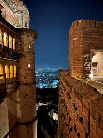 Scorcio notturno di un antico forte in pietra illuminato da luci calde; tra due alte mura si apre una vista sulla città sottostante, che brilla di luci nella notte