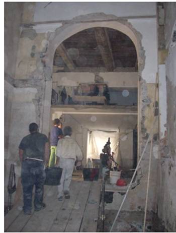 Interno di un edificio storico durante lavori di ristrutturazione: tre operai camminano su una passerella in legno verso una grande apertura muraria ad arco parzialmente demolita, con ponteggi e materiali da costruzione sullo sfondo.