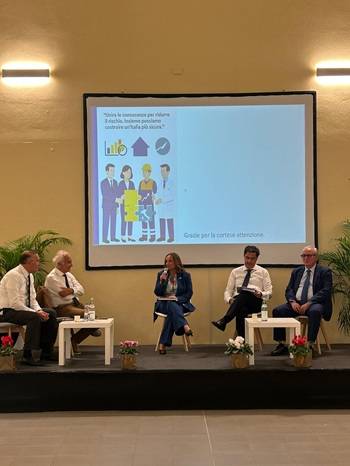 Un gruppo di cinque persone è seduto su un palco durante una conferenza o tavola rotonda. Al centro c’è una donna con microfono che parla, affiancata da quattro uomini in giacca e cravatta. Dietro di loro si vede uno schermo con una presentazione: a sinistra è visibile un’illustrazione con figure di lavoratori e professionisti, simboli di casa e lavoro di squadra; a destra appare la scritta in italiano “Grazie per la cortese attenzione”. Sul palco ci sono piante decorative e piccoli tavolini bianchi con bottiglie d’acqua e bicchieri.