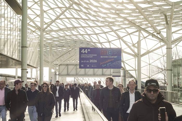 Un folto gruppo di persone cammina in una passerella coperta da una struttura in vetro e acciaio. In alto è visibile un grande striscione blu che annuncia la 44ª edizione della Mostra Convegno Expocomfort (MCE), con date dal 24 al 27 marzo 2026 e lo slogan Beyond Comfort
