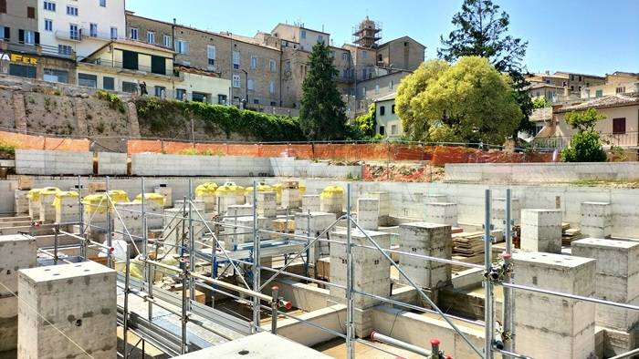 Vista panoramica di un cantiere con numerosi plinti in cemento armato e impalcature metalliche, sullo sfondo edifici residenziali e alberi.