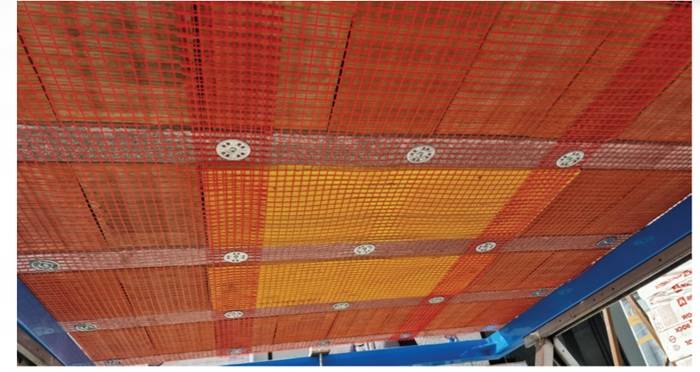 Vista dal basso di un soffitto con rete antisfondellamento installata. Le reti rosse e arancioni sono fissate tramite dischi metallici circolari distribuiti uniformemente tra i travetti in laterocemento.
