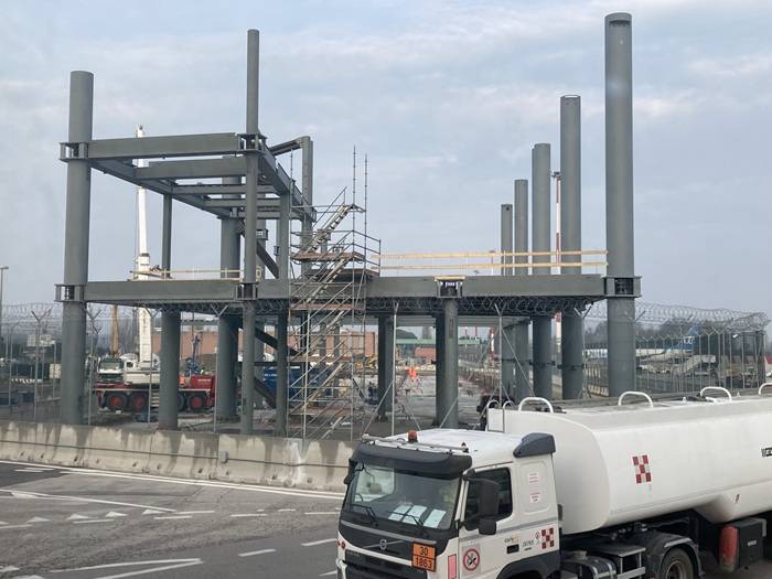 Struttura in acciaio di un edificio in costruzione con pilastri e travi parzialmente montati, ponteggi metallici e scala provvisoria al centro. In primo piano, un camion cisterna bianco è parcheggiato davanti a barriere di cemento, con recinzione e filo spinato attorno al cantiere