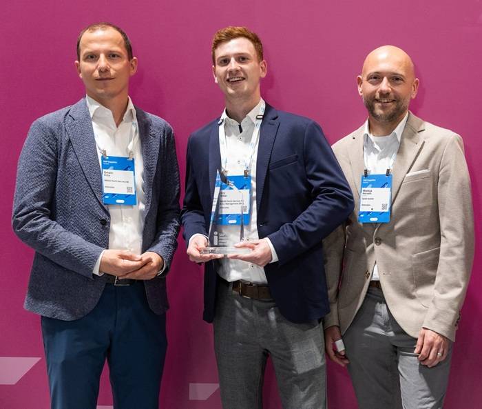 Tre uomini in abiti da business posano sorridenti davanti a uno sfondo fucsia. La persona al centro tiene in mano un premio di vetro trasparente. Tutti indossano badge identificativi da conferenza