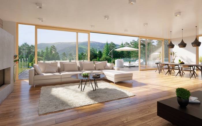 Interno luminoso di un salotto moderno con pavimento in legno, divano angolare chiaro, tavolini bassi e grandi finestre a tutta altezza che si affacciano su una terrazza con ombrellone e vista sulle montagne.