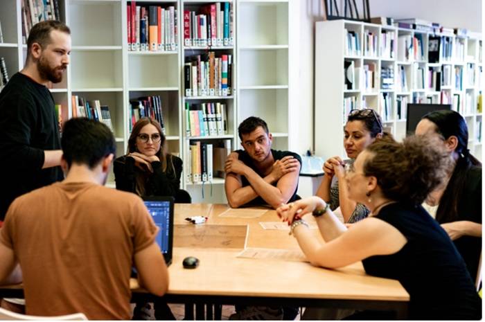 Un gruppo di persone sedute attorno a un tavolo in una biblioteca, impegnate in una discussione di gruppo. Sullo sfondo scaffali pieni di libri