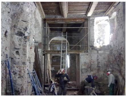 Interno di un edificio storico con murature in pietra, ponteggi metallici e operai impegnati in lavori di consolidamento attorno a un grande vano rettangolare provvisto di struttura metallica temporanea.