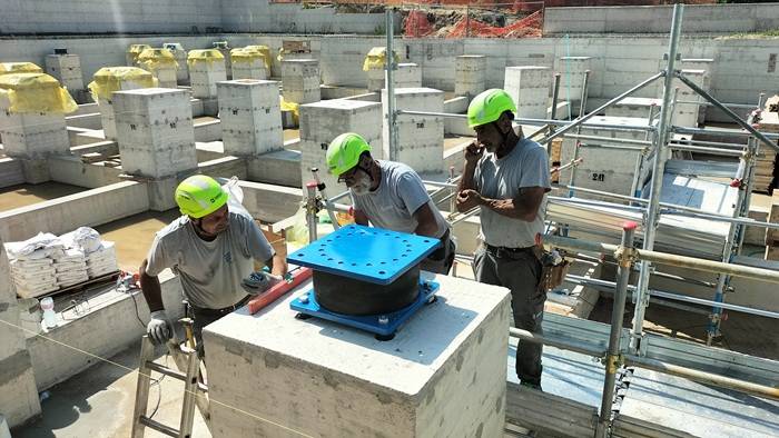 Tre operai con caschi di sicurezza giallo-verdi lavorano su un cantiere edile, installando un dispositivo antisismico blu sopra una base in cemento.