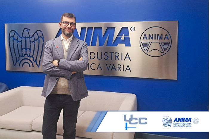 Uomo in giacca e camicia con le braccia conserte, in piedi davanti a una parete blu con il logo ANIMA Confindustria. Sullo sfondo una targa con la scritta “ANIMA – Industria Meccanica Varia” e, in primo piano, un divano chiaro; in basso a destra sono visibili i loghi UCC e ANIMA.