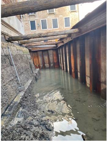 Vista dall’interno di uno scavo sostenuto da palancole metalliche arrugginite e puntellamenti in legno. Il fondo è parzialmente allagato e mostra fango e detriti; sulla sinistra è visibile un muro in pietra e laterizio.