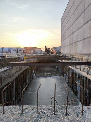 Vista di un cantiere al tramonto con una platea di fondazione in calcestruzzo appena gettata, circondata da pali di sostegno. Barre d'armatura verticali sporgono dalla superficie e una trave temporanea attraversa lo scavo.