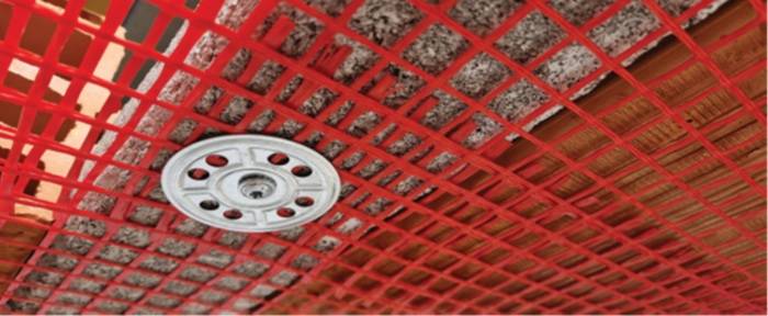 Dettaglio ravvicinato di una rete di protezione rossa fissata a un soffitto in laterocemento tramite disco metallico. Si vedono chiaramente i materiali del solaio e la trama della rete.