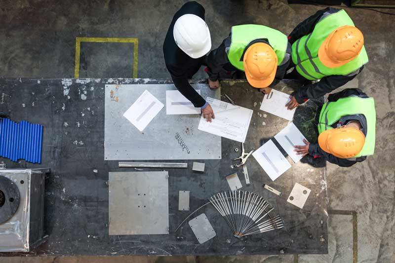 Operai con caschi e giubbotti ad alta visibilità consultano documenti tecnici su banco da lavoro industriale.